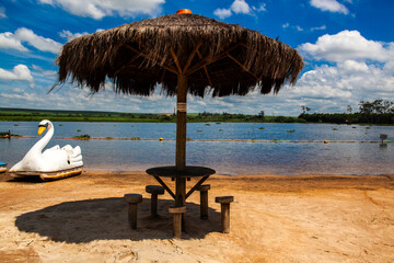 Picturesque lakeside scene with a wooden table and a thatched umbrella on a sandy beach, a swan-shaped pedal boat, calm waters, and blue sky in Brazil. Perfect for tourism, leisure, or nature themes.
