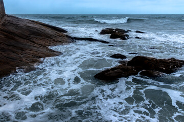 Rocky coastline with crashing waves and resilient vegetation in Brazil. A solitary island in the background adds to the dramatic coastal scene.
