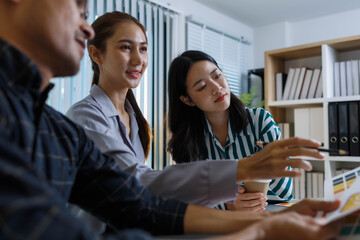 Three people are sitting at a table, working on a project. One of them is holding a piece of paper with a design on it. Scene is collaborative and focused
