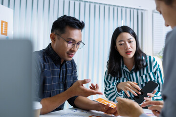 Three people are sitting at a table, working on a project. One of them is holding a piece of paper with a design on it. Scene is collaborative and focused
