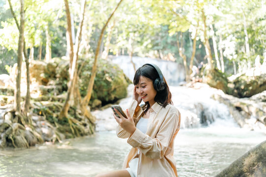 Young Asian woman listening to the music on headphones and mobile phone while travel nature hiking in forest on summer holiday vacation. People enjoy outdoor lifestyle relaxing by nature and waterfall