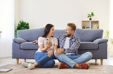 Obraz premium Happy, loving married couple shopping online together. Smiling woman holds a mobile phone and shows her husband something she wants to buy in the Internet store application