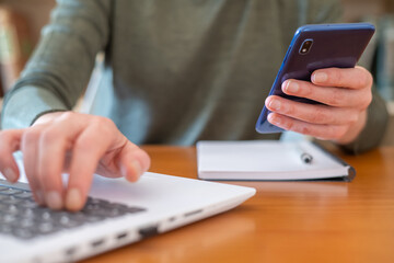 Person working on a laptop computer while holding and using a smartphone, managing multiple digital tasks and responsibilities at a wooden desk with a notebook
