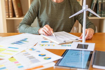 Woman managing sustainable business data, reviewing charts and a wind turbine model while focusing on renewable energy project analysis and eco-friendly technology solutions