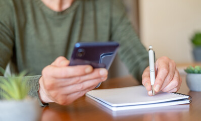 Woman's hands using a mobile phone for information while simultaneously writing notes in a spiral notebook, showcasing concepts of planning, organization, and daily task management at a desk