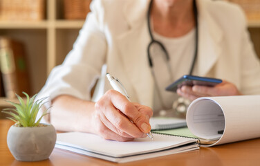 Doctor writing a prescription or medical notes during a general practitioner consultation, with a stethoscope draped around the neck and holding a cell phone, representing modern healthcare practice
