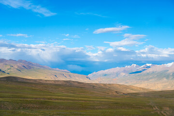 Expansive view of a vast, high-altitude grassland and rolling hills under a bright blue sky with snowy mountain peaks in the distance