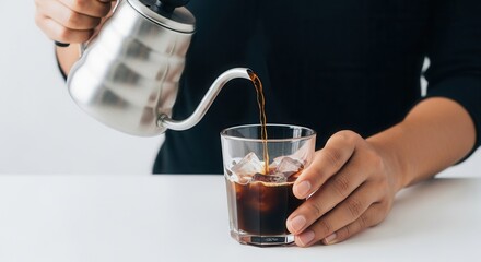 A person in a dark shirt pours a dark liquid from a silver gooseneck kettle into a glass with ice cubes and liquid on a white surface
