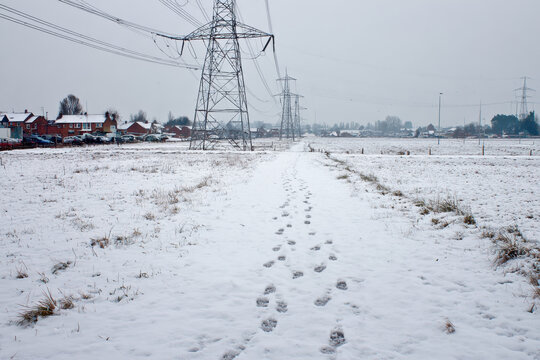 Snowy landscape with footprints leading through field, featuring power lines and houses in background under cloudy sky. scene conveys sense of calm and solitude