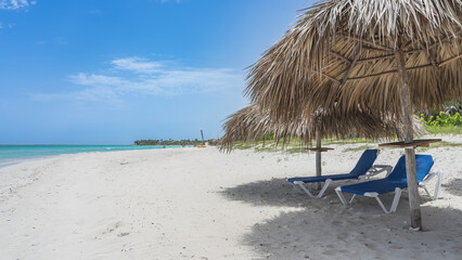 Two sun loungers on the sandy beach in the shade of straw umbrellas. Calm aquamarine ocean, blue sky, clouds. Cuba. Varadero. Resort. Hotel.