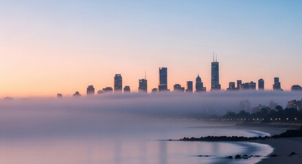 Obraz premium Skyline of tall structures rising above a thick fog layer reflecting on calm coastal waters under a pastel gradient sky with a visible shoreline