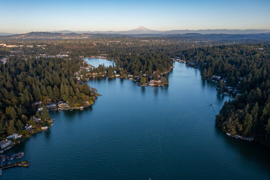 Aerial drone picture of Lake Oswego, Oregon and Mount Hood in backgrond, showing calm blue water surrounded by dense forest and residential houses, real estate, on a clear sunny evening at sunset.  