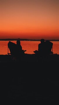 Silhouette of couple in chairs watching vibrant sunset, calm lake