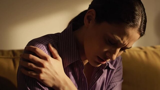 Woman in Purple Shirt with Shoulder Pain in a Sun Drenched Room with Light Brown Wall and Sofa Feeling Aching and Discomfort Showing Suffering and Distress in Warm Lighting