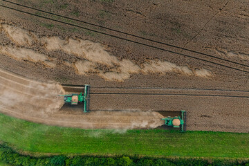 Two green combine harvesters work side by side in a large field, bordered by green grass and trees, leaving parallel tracks and trails of dust.