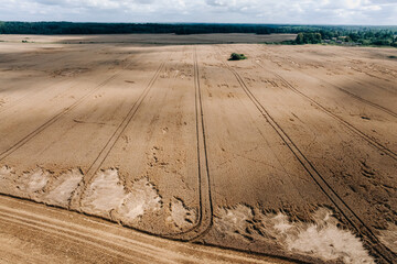 A vast golden field with parallel lines from machinery, patches of flattened crops, a forested border, scattered structures, and a partly cloudy sky.