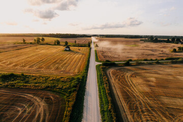 A straight dirt road runs through golden fields with visible crop patterns, a small structure on the left, and trees lining the horizon under a cloudy sky.