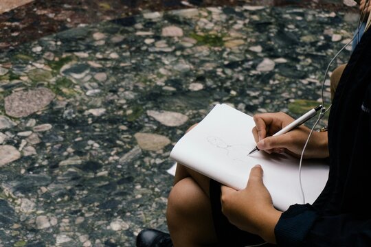 Woman drawing (or illustrating) in book while sitting down, listening to music using wired apple earphones