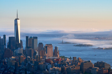 Aerial view of Manhattan skyline with One World Trade Center and Statue of Liberty emerging from morning fog over New York Harbor.