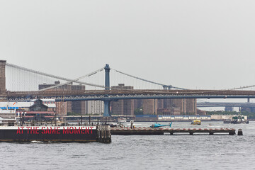 View of a helicopter landing pad on the East River in Lower Manhattan, New York City, with Brooklyn Bridge and Manhattan Bridge in the background. Urban skyline and river traffic visible.
