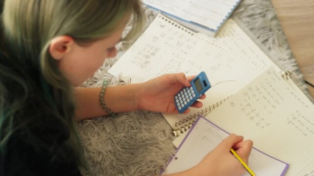 Teenage girl lying on cozy carpet while do math homework, diligently writing in notebook and using small calculator to solve complex problems and equations for her studies