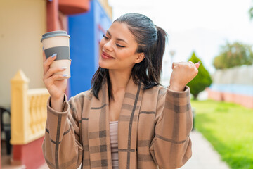 Young pretty woman holding a take away coffee at outdoors celebrating a victory
