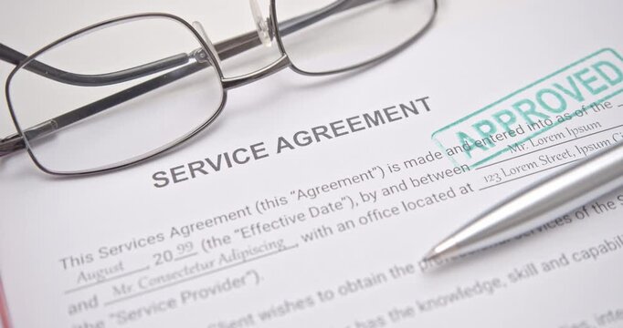 Green APPROVED stamp on service agreement paper beside pen and eyeglasses, representing verified legal document, successful business contract, and approval process.