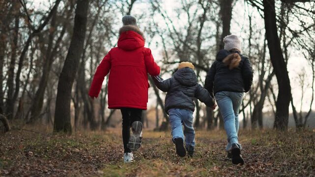 Child walks through winter forest path with kid sister and brother holding hands, family play and walk on park trail, child and sibling running together under bare tree canopy while laughing