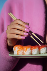 Woman with glossy pink nails holding sushi with chopsticks against a bright background. Contemporary food styling with color-coordinated fashion and minimalism.