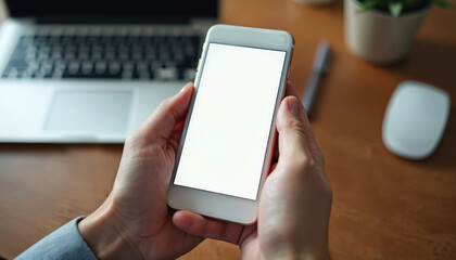 Man holds white smartphone with empty screen at office desk. Laptop, mouse in background. Person uses mobile device for work, business, connection, or online activity in modern environment.