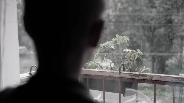 The boy stands at the window behind which there is a downpour and looks sadly at the street. He's in a sad mood from the weather.