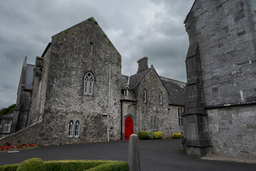 The Trinitarian Abbey in Adare, Ireland, features a striking red door and gothic architecture, set against a cloudy sky, surrounded by lush greenery
