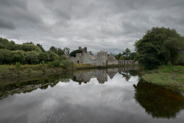 The Desmond Castle ruins reflected in the calm waters of the River Maigue under an overcast sky, surrounded by lush greenery, Adare, Ireland