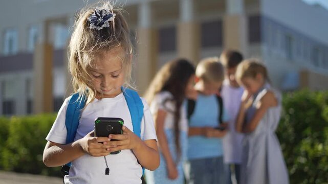 Girl using smartphone at school focused on screen while child peer and group behind whisper and point showing bullying and isolation backpack and playground visible with texting and closed posture