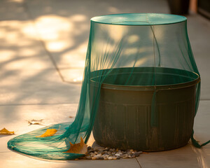 Close up of a green mesh cover over a dark green container with leaves and stones on a tiled surface