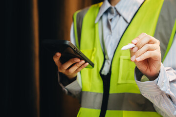 Construction Worker in Safety Vest Using Smartphone and Holding Pen for Notes in Industrial Environment
