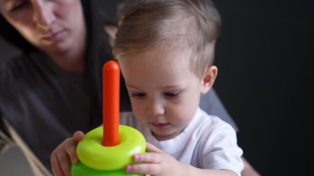 Toddler plays with stacking ring toy while parent guides hand as child explores stacker toy learning color and shape through play with mother and baby promoting development and focus and concentration