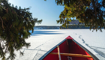 A red canoe rests on the edge of a frozen, snow-covered lake on a sunny winter day, framed by evergreen branches