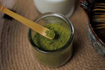 A wooden spoon holds a scoop of bright green matcha powder over a glass container filled with more powder.