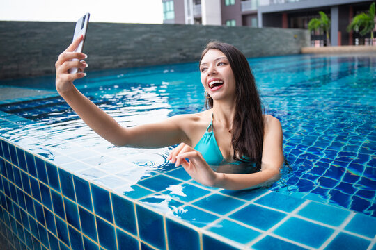 Young Hispanic Gen Z Woman Using Smartphone to Update Social Media by Resort Pool During Summer Vacation.
