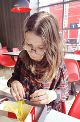 Young girl in glasses sits in a cafe and eats French fries. Delicious unhealthy food.