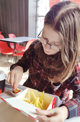 Young girl in glasses sits in a cafe and eats French fries. Delicious unhealthy food.