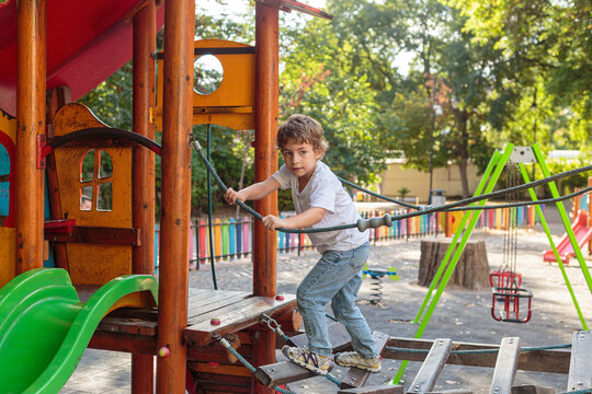 Active little boy climbing a wooden rope bridge on a colorful playground structure in a sunny park during a summer afternoon