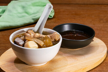 Fish maw soup in white ceramic bowl with white ceramic spoon and seasoning sauce in black ceramic cup on wooden board on wooden table.