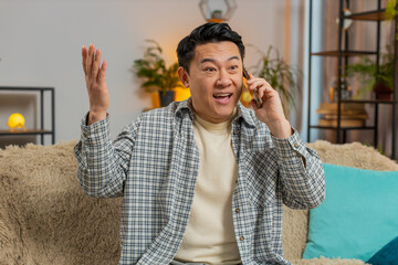 Smiling young adult Chinese man making fun phone conversation with friends sitting on couch sofa at home in living room. Happy excited guy Middle Eastern salesman enjoying mobile talking indoors.