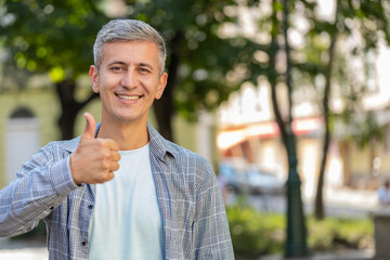 Fototapeta premium Like. Happy mature man raises thumbs up agrees with something gives positive reply recommends advertisement good idea feedback, celebrating success victory outdoors. Middle-aged guy on city street.