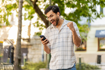 Indian young man using mobile smartphone celebrating win good message news, lottery casino jackpot victory, giveaway online outdoors. Hispanic guy on urban city street. Town lifestyles. Horizontal.