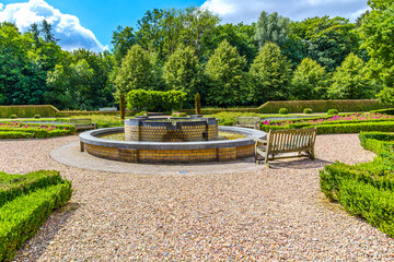 Fountain between pedestrian paths, green bushes and pink flowers, public park in front of TerWorm Castle, surrounded by leafy trees, sunny spring day in Heerlen, South Limburg, Netherlands