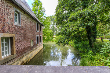 Moat surrounding TerWorm Castle seen from entrance bridge, red brick walls and windows, surrounded by leafy trees, cloudy spring day in Heerlen, South Limburg, the Netherlands