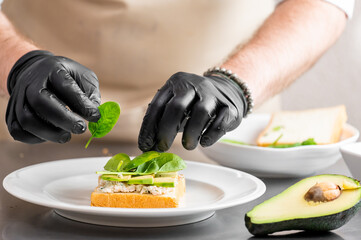 Person in gloves preparing healthy open-faced sandwich with avocado, spinach, and salad spread on white bread. Fresh ingredients and clean kitchen setup. Top view, food styling